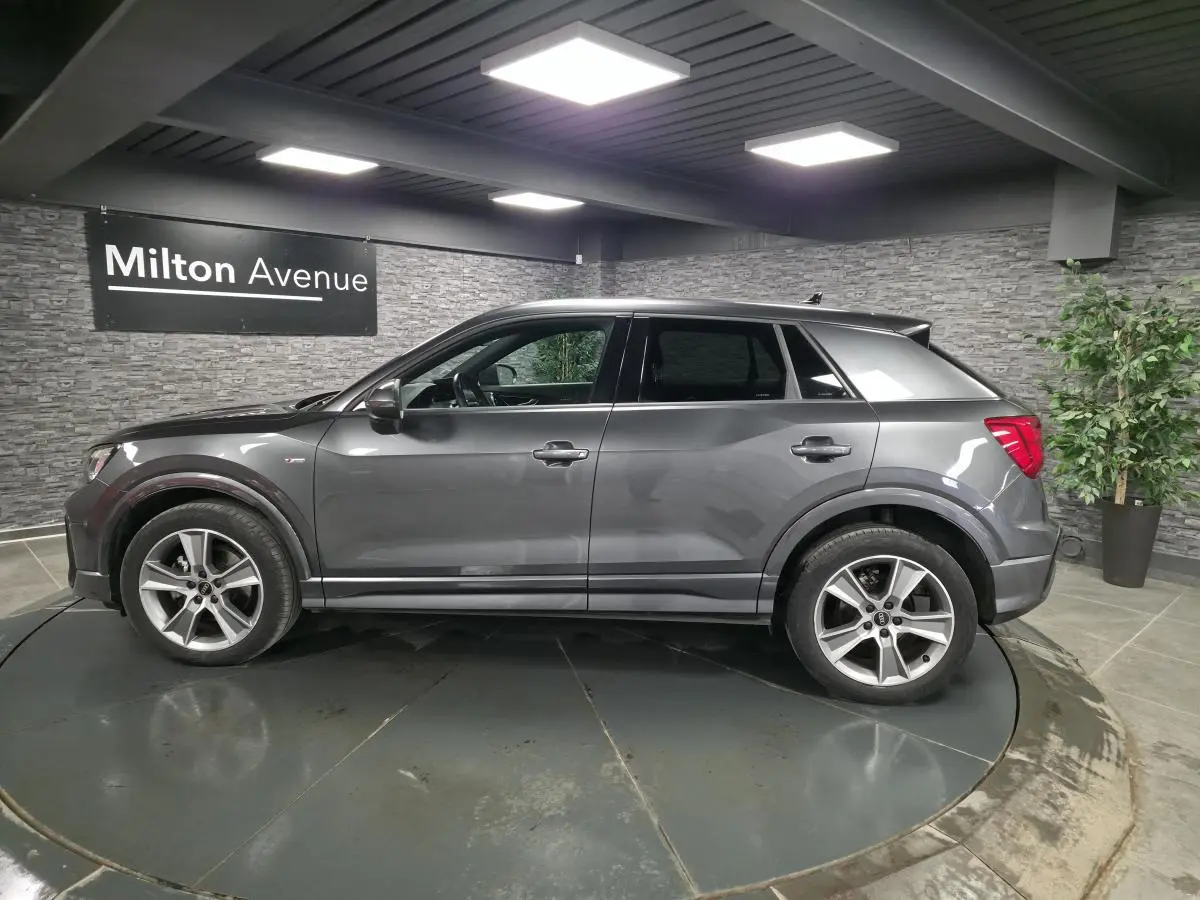 Audi Q2 gris Daytona en vue profil droit dans un showroom avec jantes alliage 18 pouces et vitres surteintées.