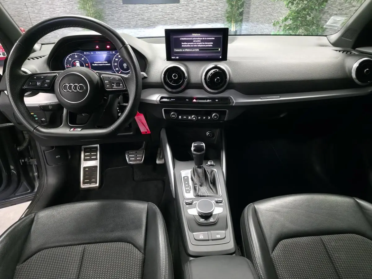 Vue intérieure avant de l'Audi Q2 S line Plus 2023, tableau de bord noir avec volant sport multifonction et console centrale moderne.