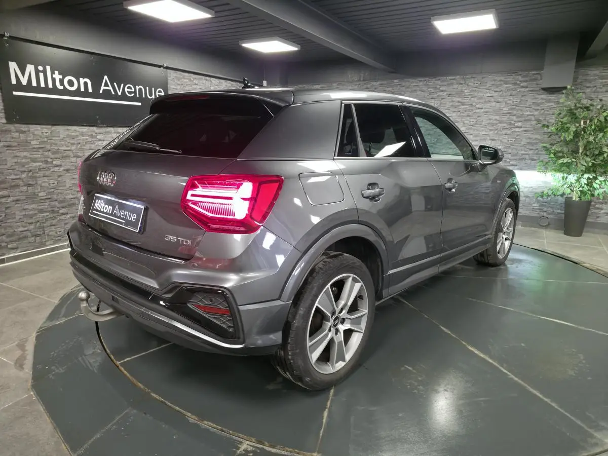 Audi Q2 gris en 3/4 arrière droit, avec feux LED allumés et attelage amovible visible au pare-chocs.