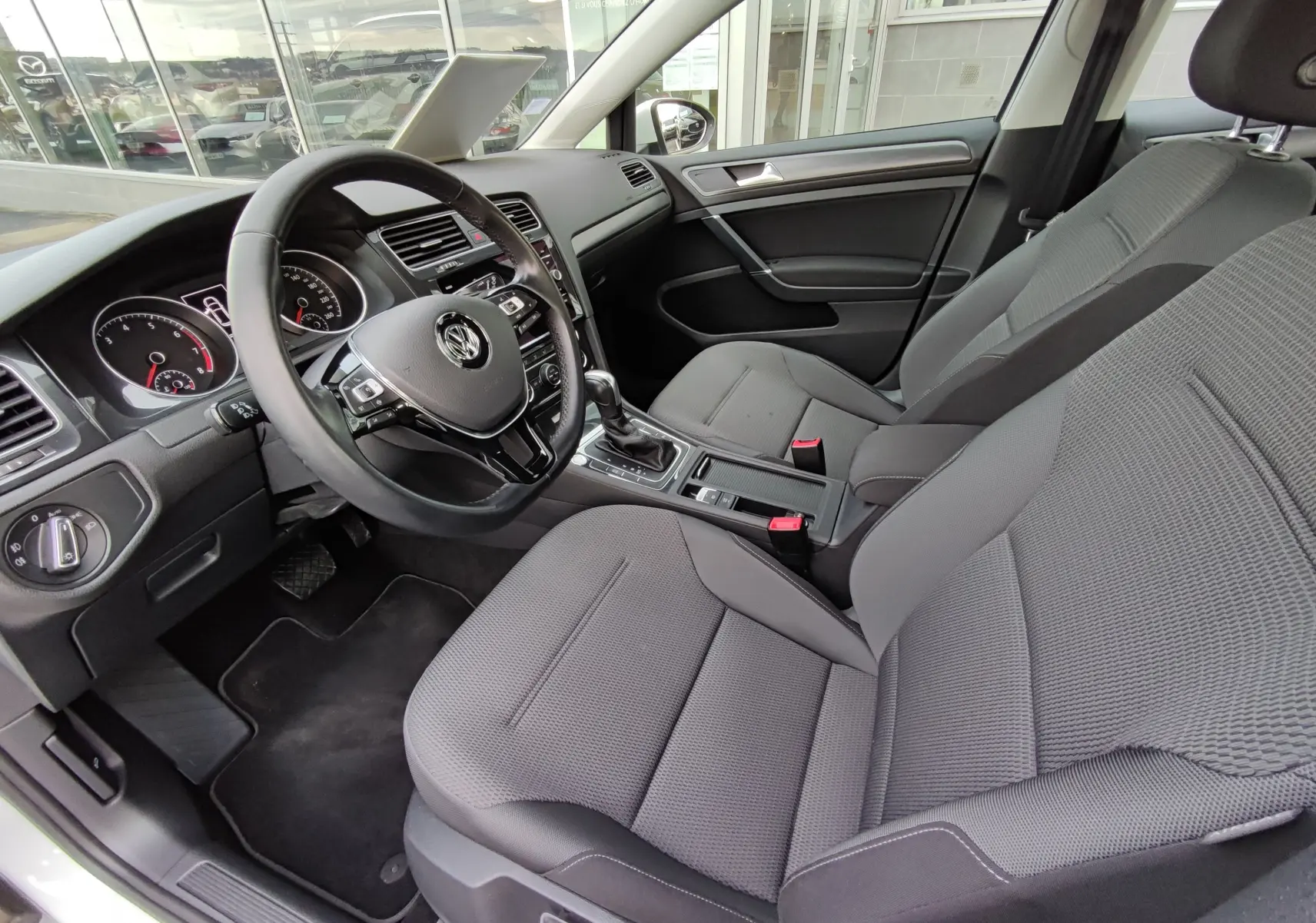 Vue intérieure côté conducteur de la Volkswagen Golf blanche 1.5 TSI Evo 2019, avec tableau de bord et sièges gris.