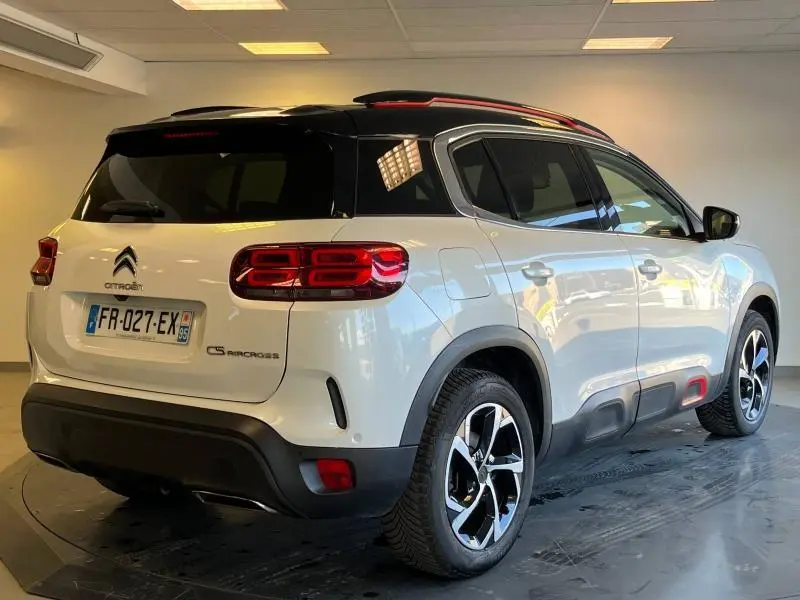 Vue 3/4 arrière droite d'un Citroën C5 Aircross blanc nacré avec toit noir et jantes alu bicolores en intérieur showroom.