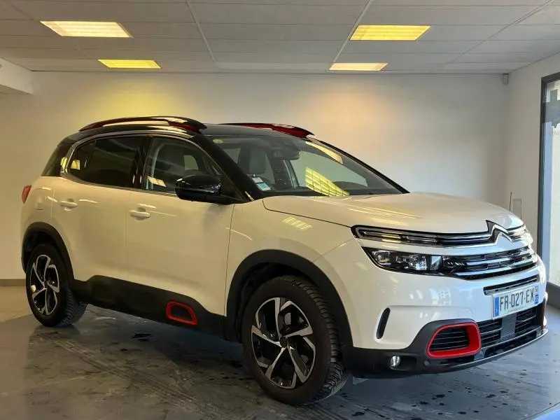 Vue 3/4 avant d'un Citroën C5 Aircross blanc nacré avec détails rouges et toit noir en intérieur showroom.