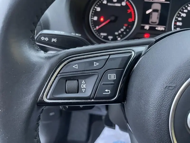 Gros plan sur les commandes gauche du volant noir de l'Audi Q2 30 TFSI 116 SPORT LIMITED 2020, avec tableau de bord partiel en arrière-plan.