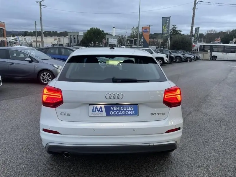 Vue arrière d'un Audi Q2 blanc 30 TFSI 116 SPORT LIMITED de 2020 avec feux allumés sur parking extérieur.