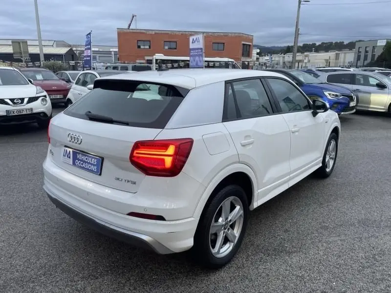 Audi Q2 blanc vue 3/4 arrière droit, feux arrière allumés, stationné sur parking extérieur.
