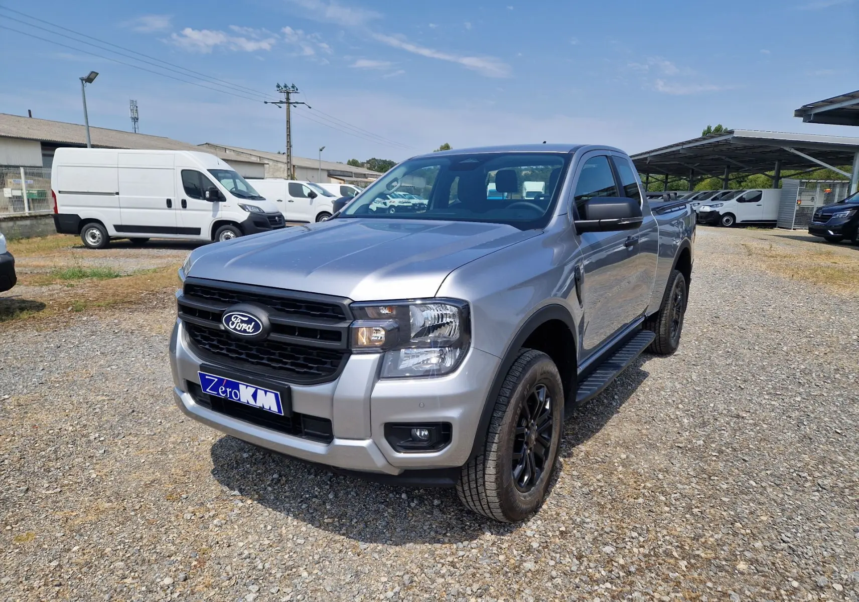Ford Ranger Super Cabine gris Iconic vu en 3/4 avant droit, avec marchepied noir et jantes foncées sur terrain gravillonné.