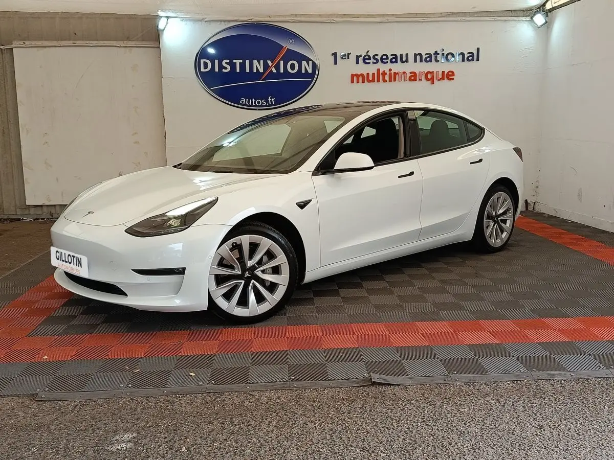 Tesla Model 3 Grande Autonomie blanc vue 3/4 avant droit, stationnée sur sol à damiers rouge et noir en intérieur.