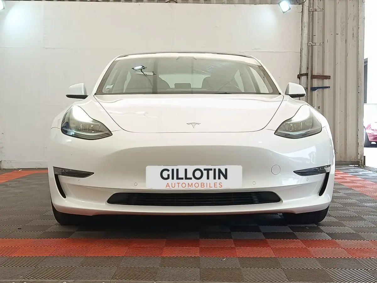 Vue frontale d'une Tesla Model 3 Grande Autonomie blanche, avec phares LED allumés et plaque d'immatriculation GILLOTIN.