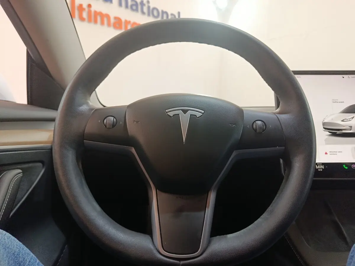 Vue rapprochée du volant en cuir synthétique noir d'une Tesla Model 3 blanche, avec écran tactile visible à droite.