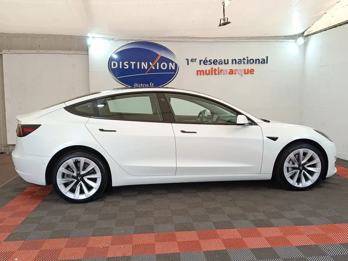 Tesla Model 3 Grande Autonomie blanche vue de profil côté gauche dans un showroom avec sol à damiers rouge et gris.