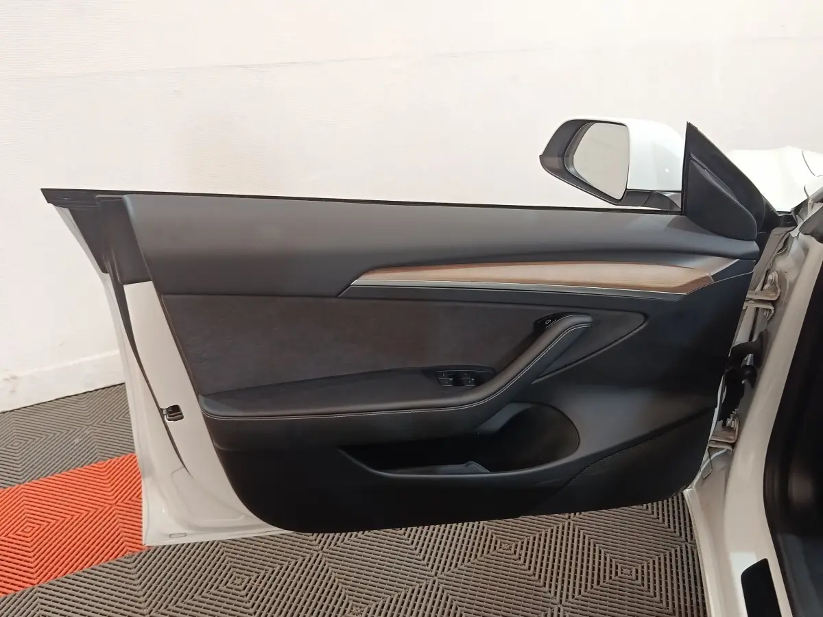 Porte avant gauche ouverte d'une Tesla Model 3 blanche, intérieur noir avec insert bois et commandes électriques.