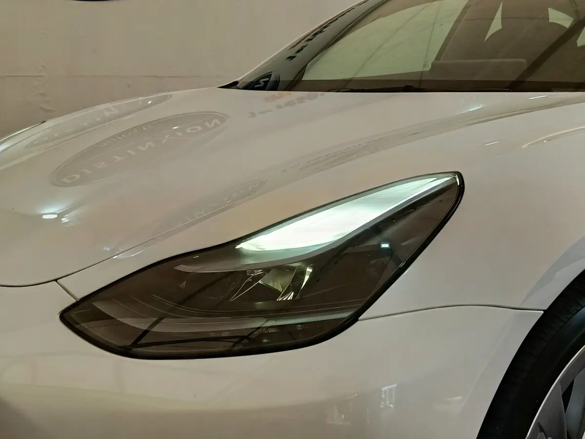 Gros plan sur le phare avant gauche d’une Tesla Model 3 blanche, mettant en valeur son design épuré et moderne.