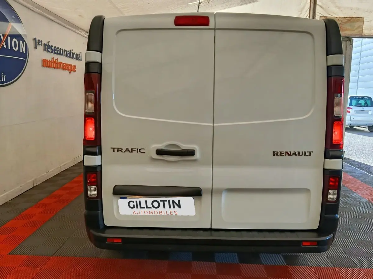 Vue arrière d’un Renault Trafic fourgon blanc avec portes battantes et feux arrière allumés en intérieur showroom.