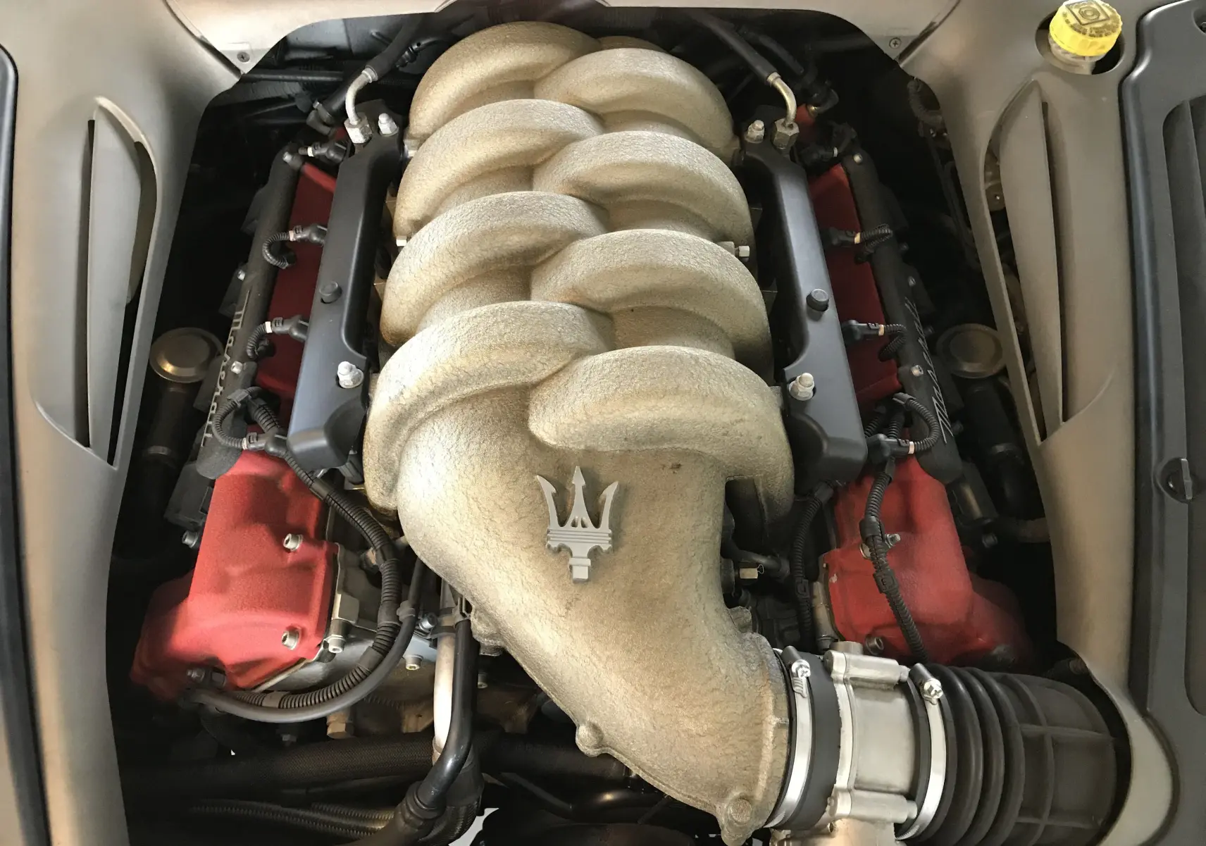Gros plan sur le moteur V8 4.2l gris clair avec logo Maserati, entouré de pièces rouges et éléments mécaniques visibles.