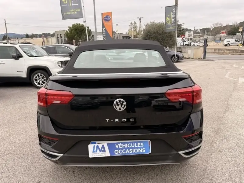 Vue arrière d’un Volkswagen T-Roc Cabriolet noir métal avec toit souple fermé sur un parking extérieur.