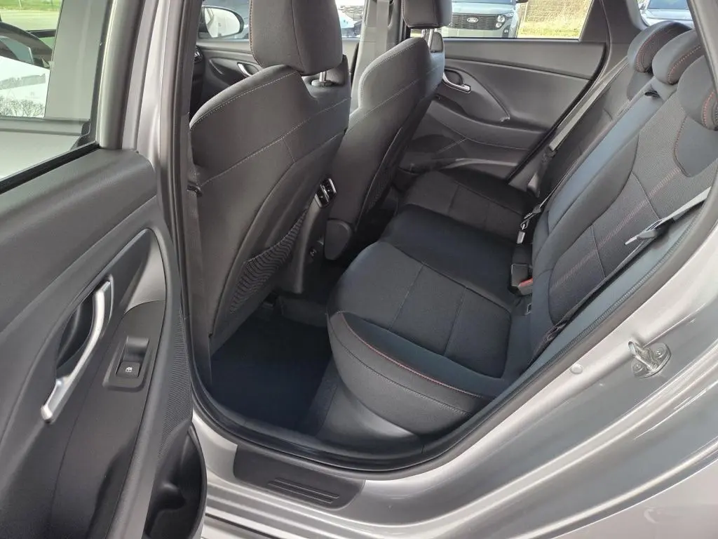 Vue intérieure côté arrière droit de la Hyundai i30 SW N-LINE 2025, sièges tissu noirs avec surpiqûres rouges, porte gris foncé ouverte.
