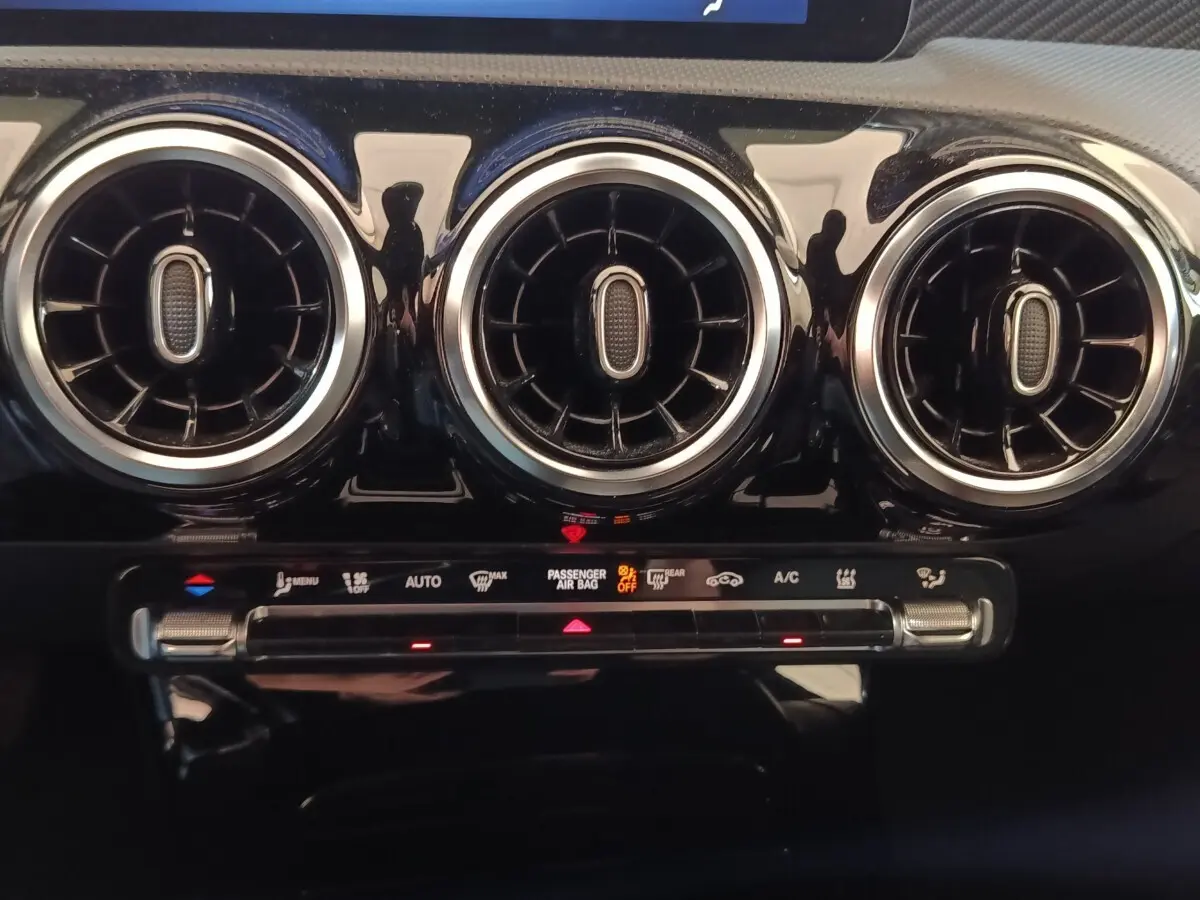 Gros plan sur les trois aérateurs ronds chromés et la commande de climatisation du tableau de bord noir brillant du Mercedes GLA 250 e.