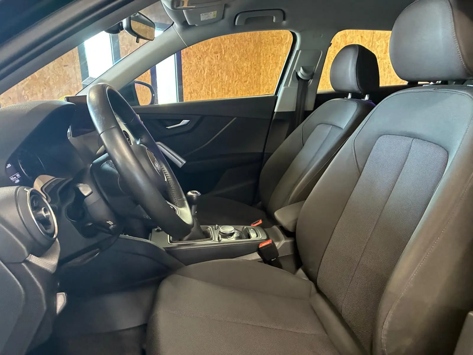 Vue intérieure côté conducteur de l'Audi Q2 noir, montrant les sièges en tissu noir et la console centrale avec boîte manuelle.
