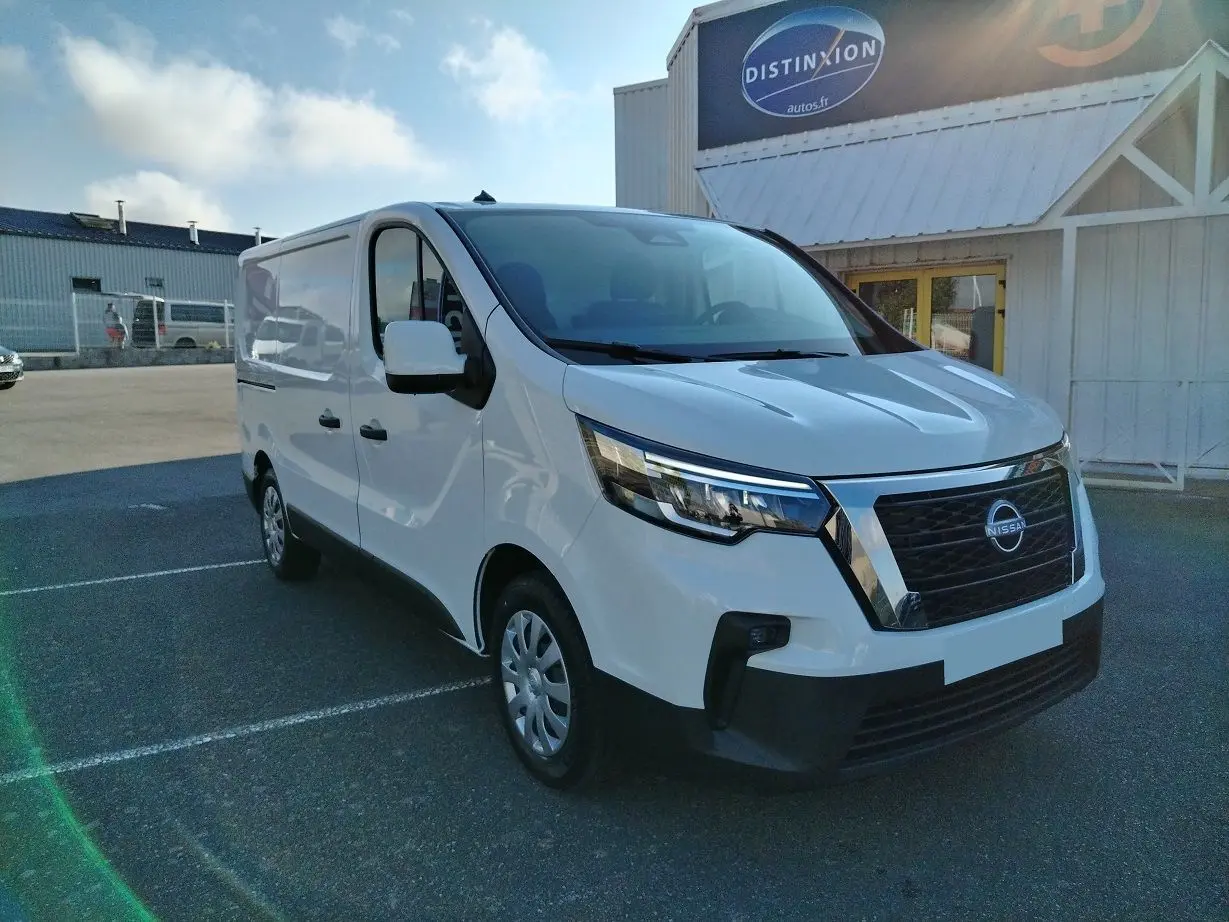 Nissan Primastar blanc en 3/4 avant droit, utilitaire avec calandre noire et détails chromés sous un ciel bleu.