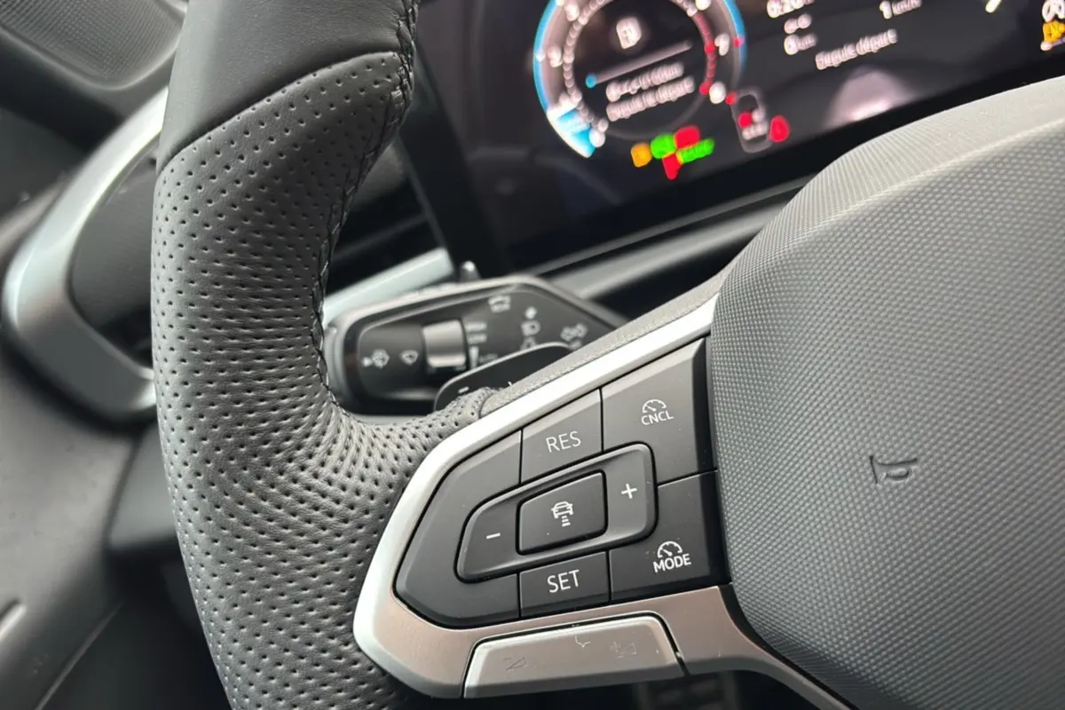 Gros plan sur le volant cuir perforé multifonctions du Volkswagen T-Roc gris loup, avec tableau de bord numérique en arrière-plan flou.