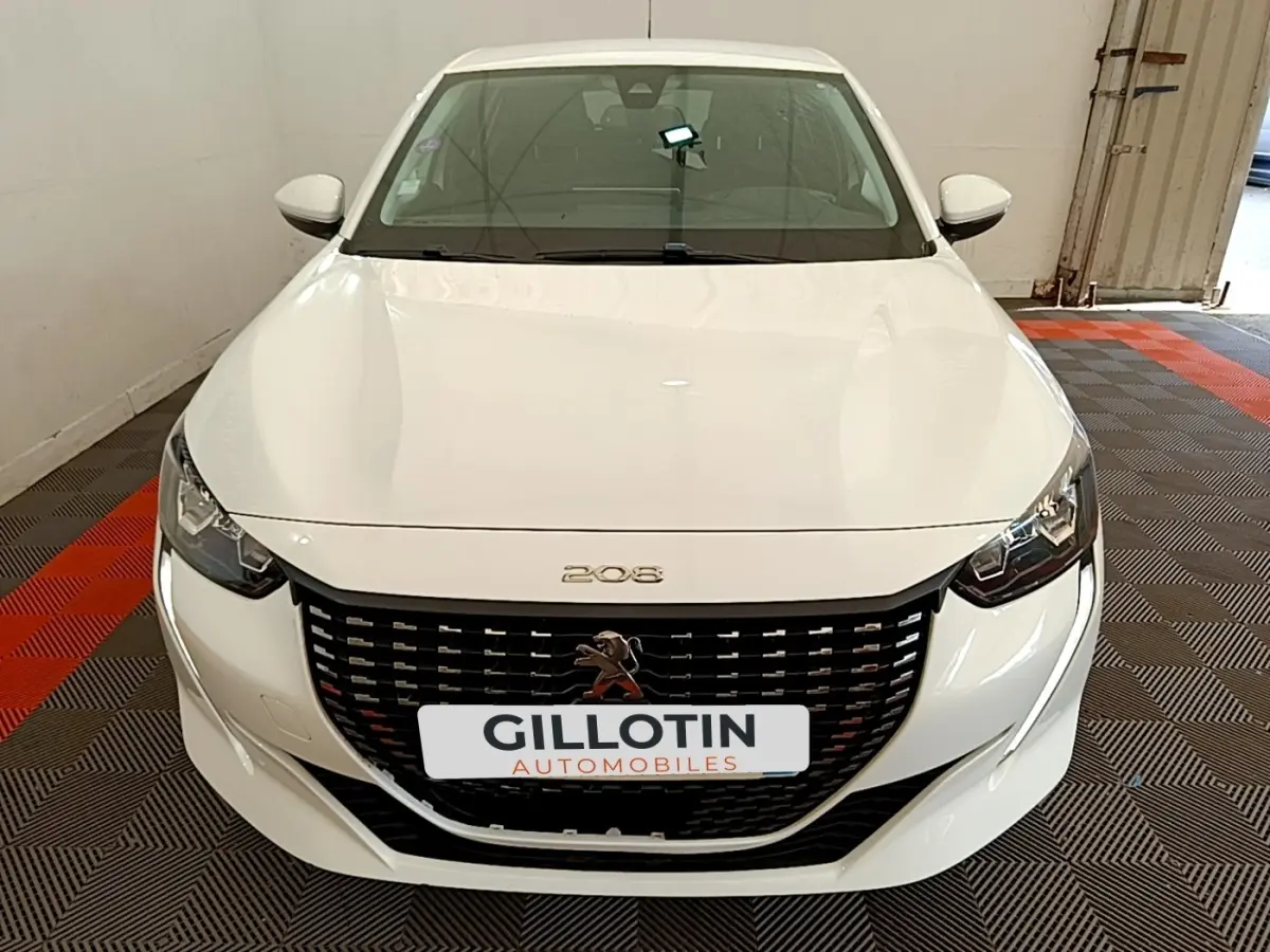 Vue avant d'une Peugeot 208 blanche avec calandre noire et logo lion au centre, stationnée en intérieur.