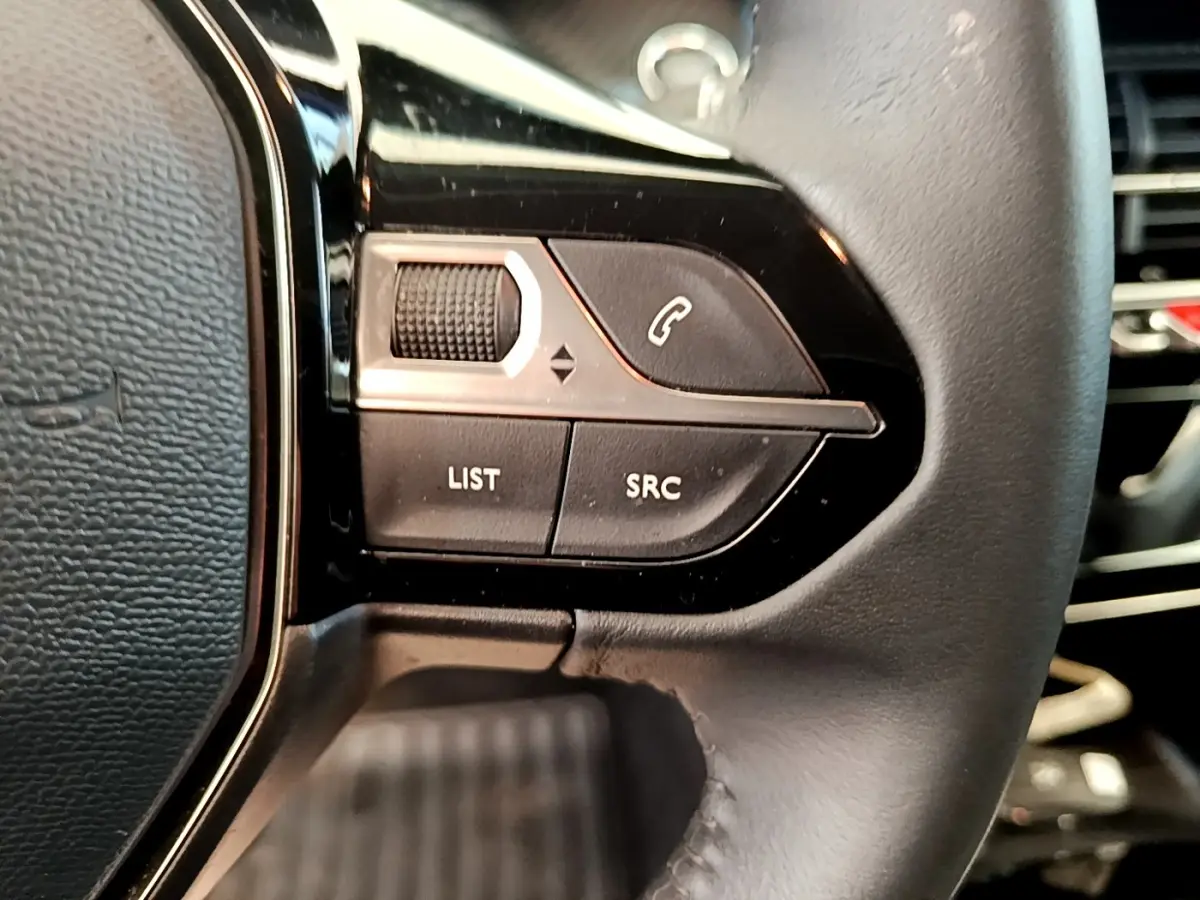 Gros plan sur les commandes gauche du volant cuir noir du Peugeot 208 blanc, avec boutons de gestion audio et appel.