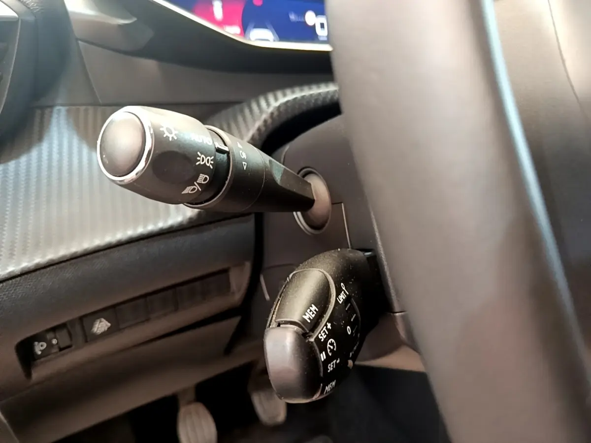 Gros plan sur la colonne de direction à gauche du volant cuir d’une Peugeot 208 blanche, avec commandes de phares et régulateur.