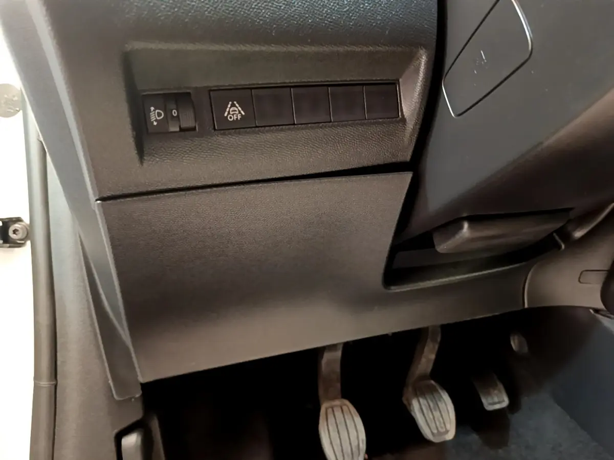 Vue rapprochée sous le tableau de bord côté gauche montrant les commandes d'éclairage et les pédales d'une Peugeot 208 blanche.