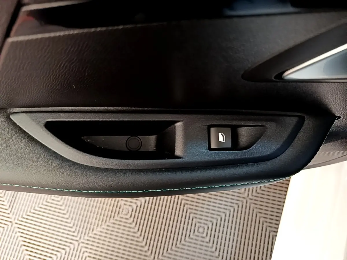 Gros plan sur la commande de lève-vitre côté conducteur d'une Peugeot 208 blanche, avec surpiqûres bleues sur la porte.