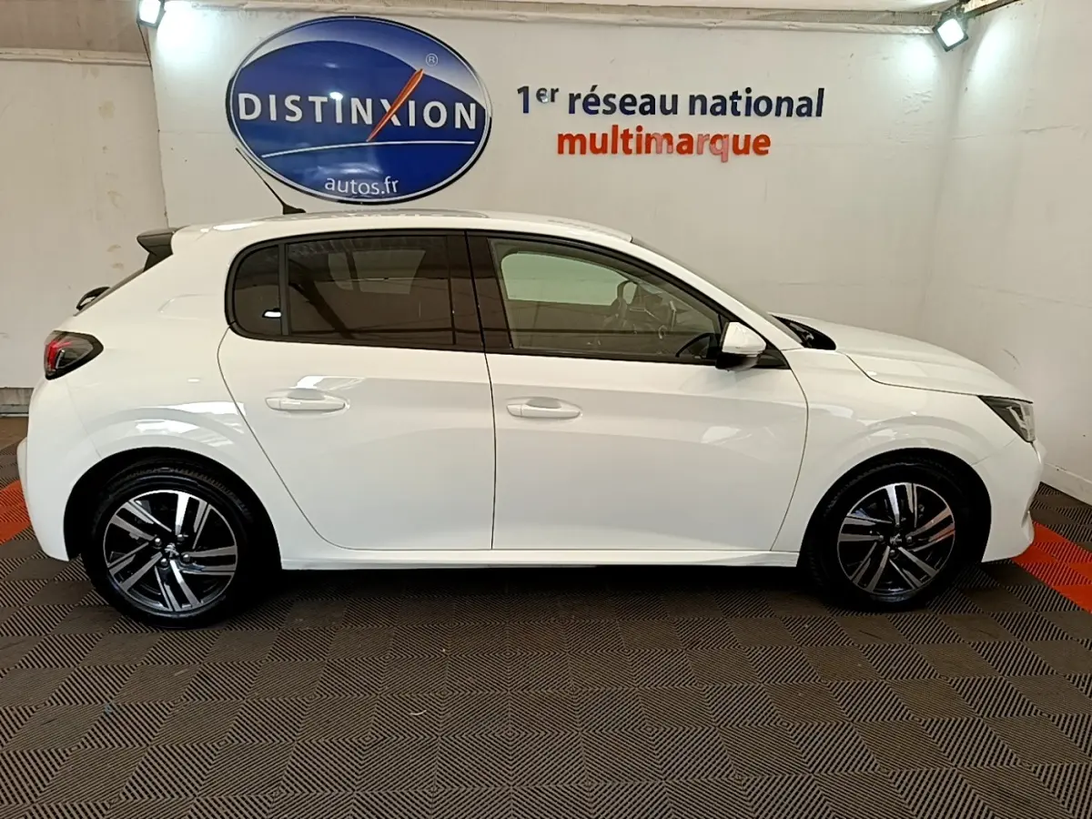 Profil droit d'une Peugeot 208 blanche de 2021, avec jantes alliage noires et vitres teintées, en intérieur showroom.
