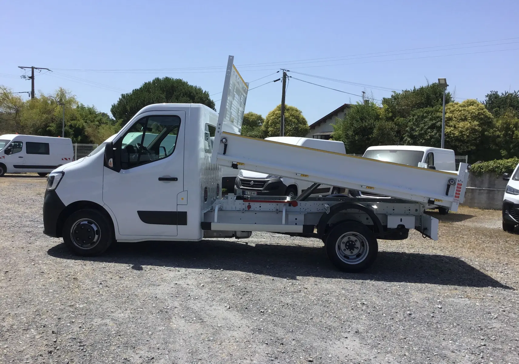 Vue de profil côté gauche d’un Renault Master blanc avec benne basculante relevée sur un terrain extérieur.