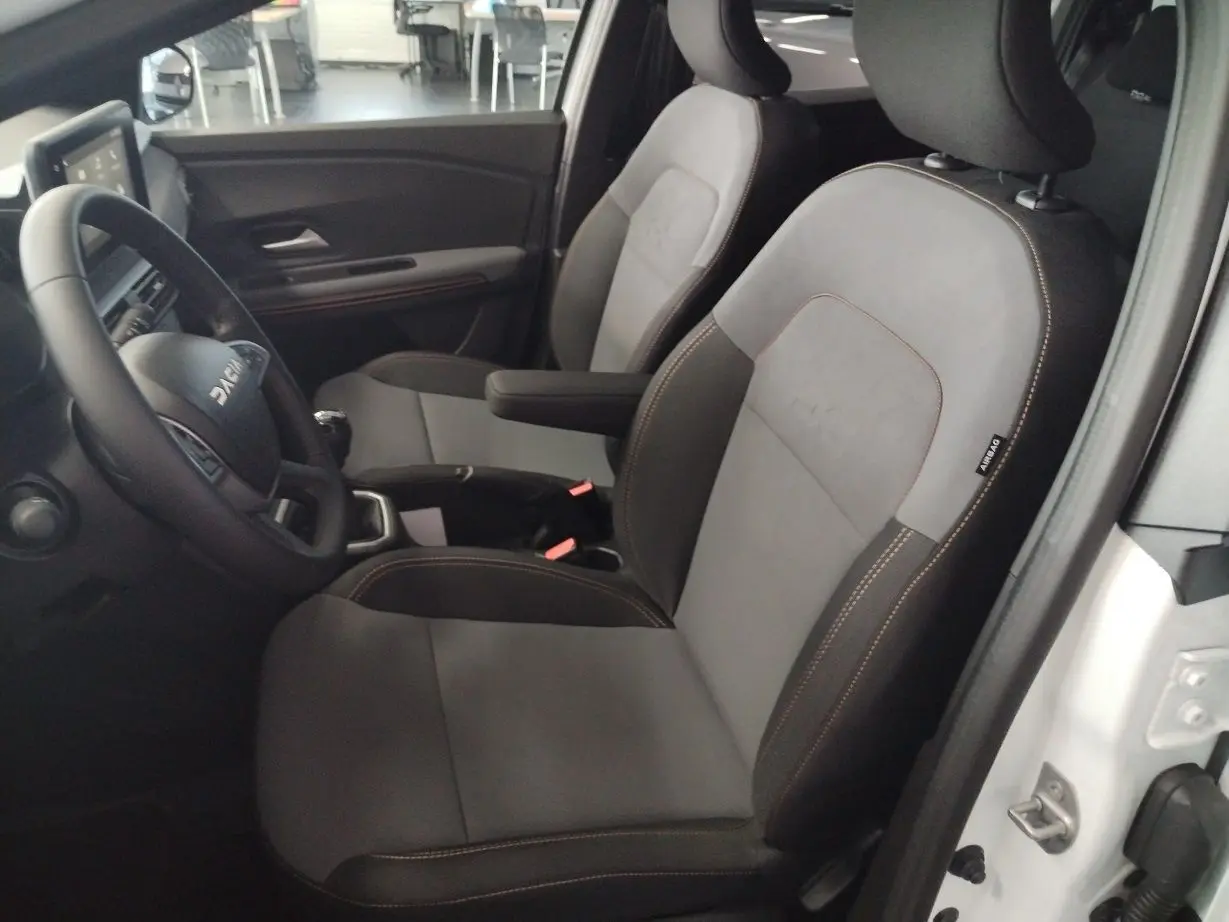 Vue intérieure côté conducteur du Dacia Jogger 2025 blanc, sièges gris avec surpiqûres marron et volant multifonction.