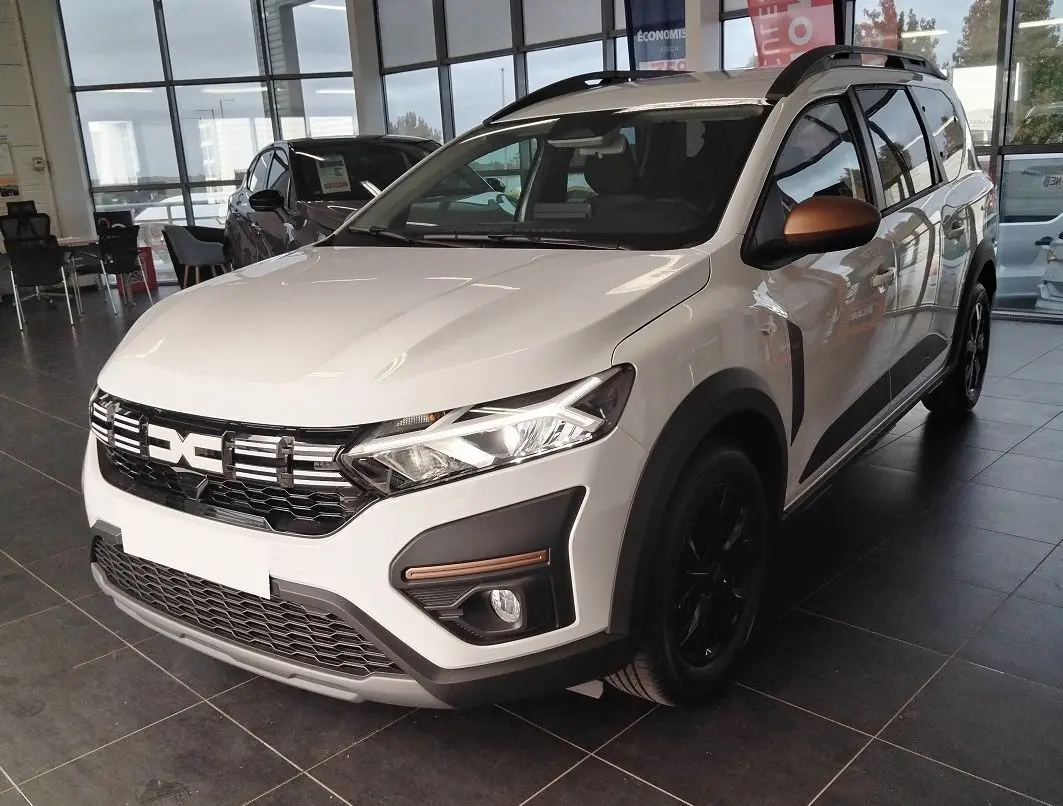 Vue 3/4 avant d'un Dacia Jogger blanc 2025 avec barres de toit modulables et rétroviseurs cuivrés en showroom.