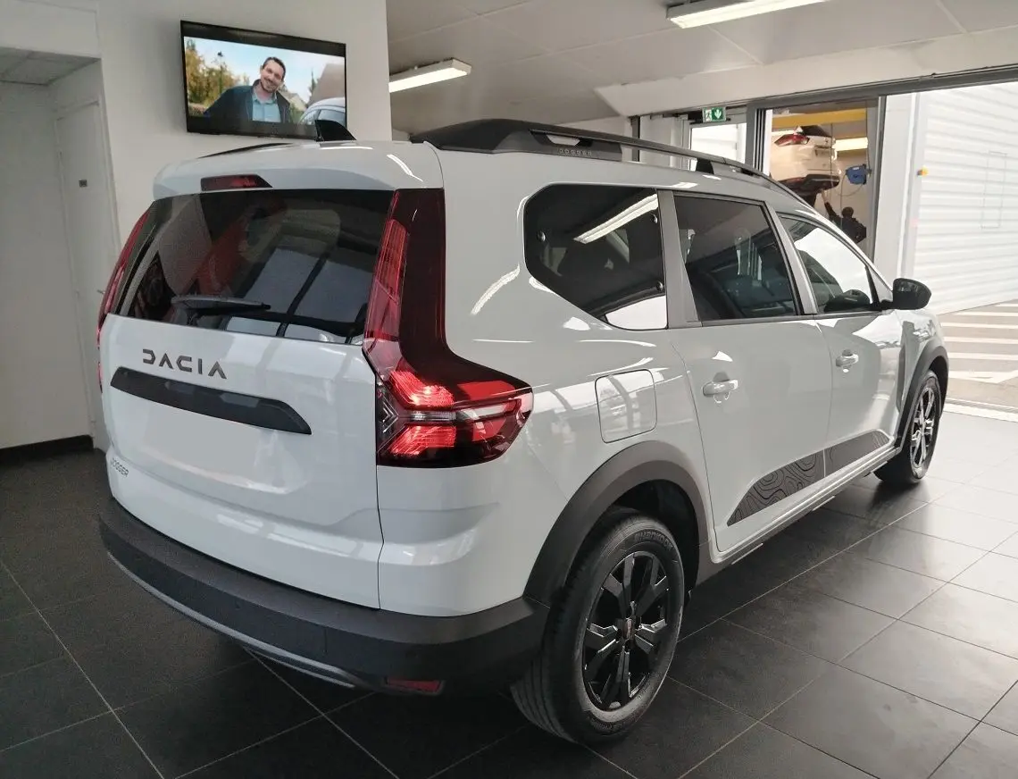 Vue 3/4 arrière droite du Dacia Jogger blanc avec barres de toit modulables et jantes noires en showroom.