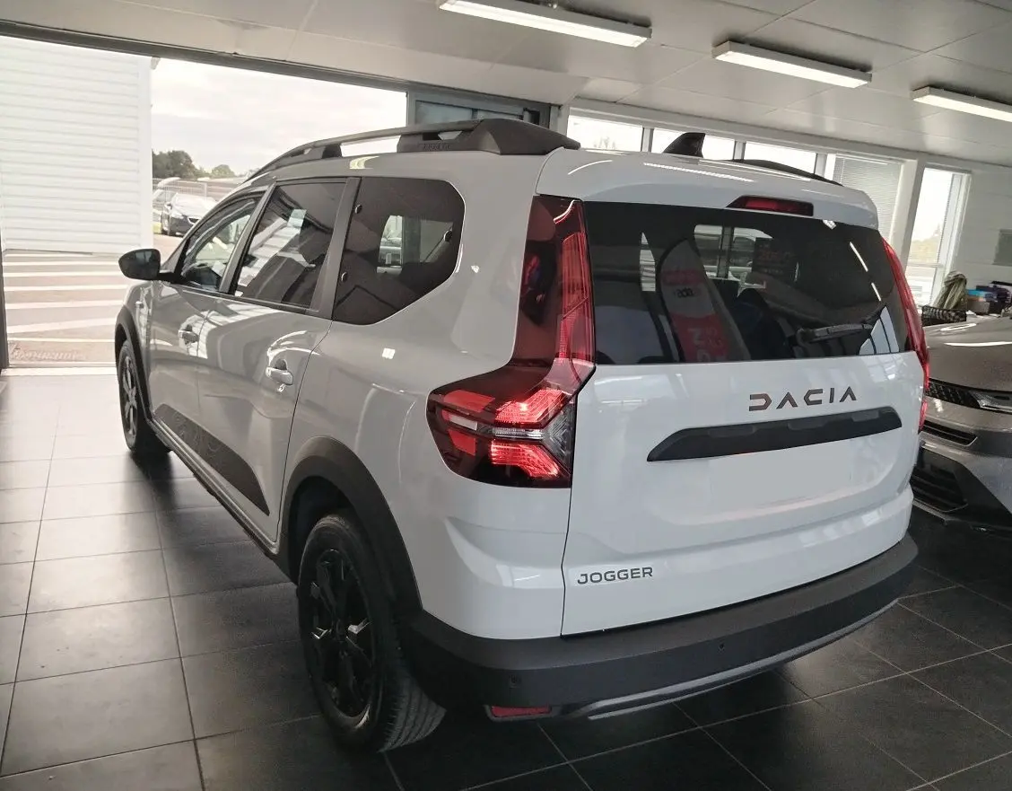 Vue 3/4 arrière d'un Dacia Jogger blanc avec barres de toit modulables et feux arrière LED en intérieur showroom.