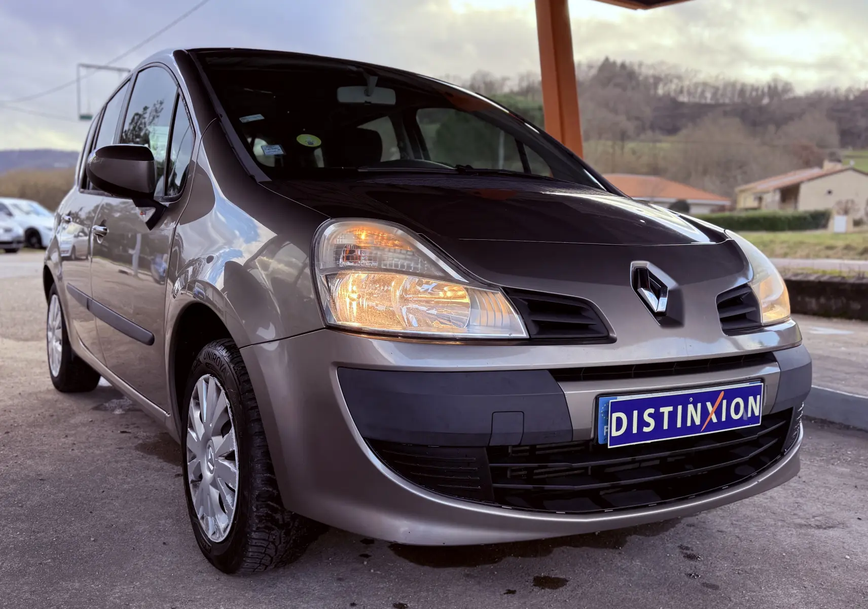 Renault Modus beige vue 3/4 avant droit avec phares allumés et plaque Distinxion visible.