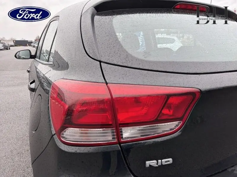 Gros plan sur le feu arrière droit d'une KIA Rio noire Ebène sous la pluie, avec le logo RIO visible.