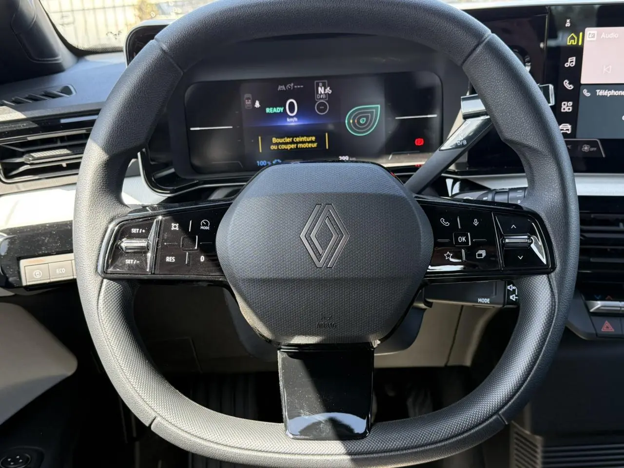 Vue rapprochée du volant noir grainé avec logo Renault, tableau de bord numérique et commandes multifonctions visibles.