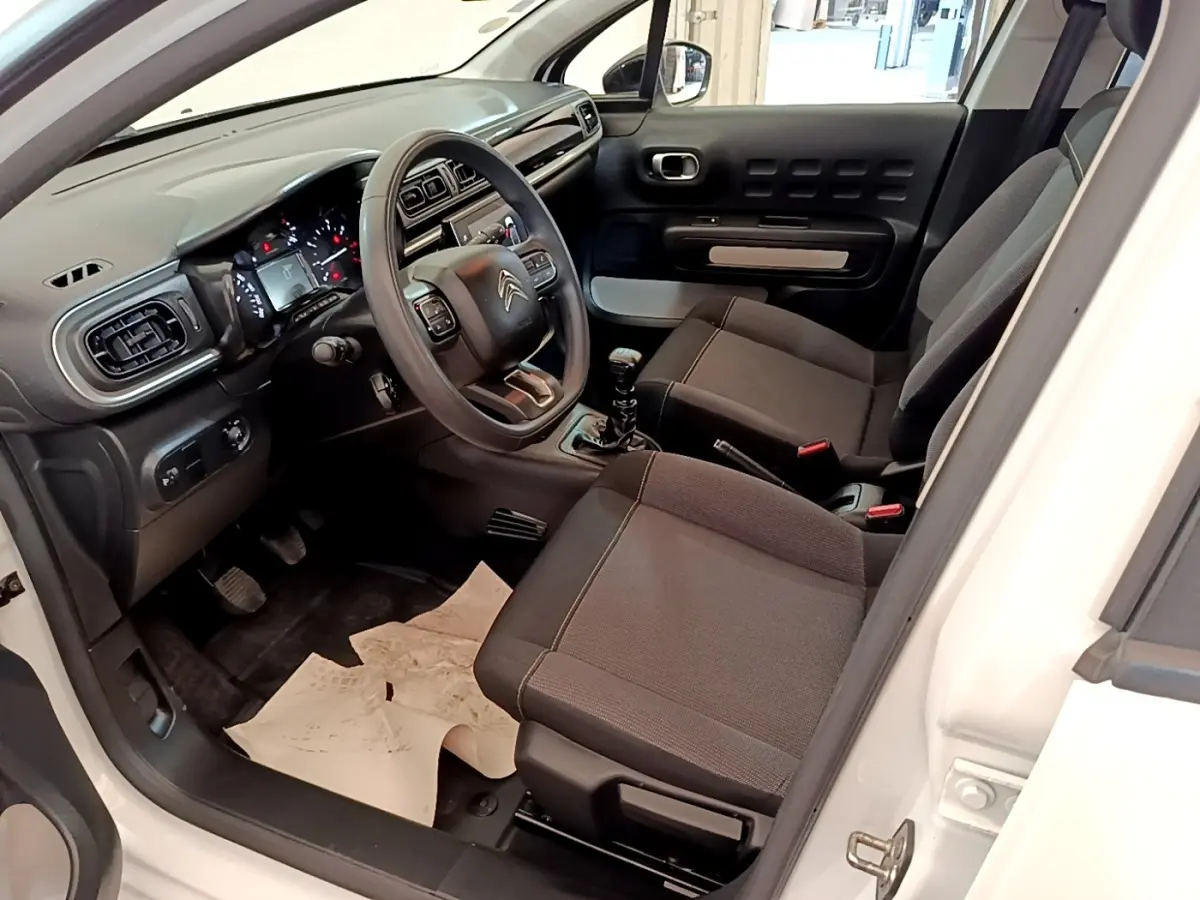 Vue intérieure côté conducteur d'une Citroën C3 blanche de 2021, montrant le tableau de bord et les sièges noirs.