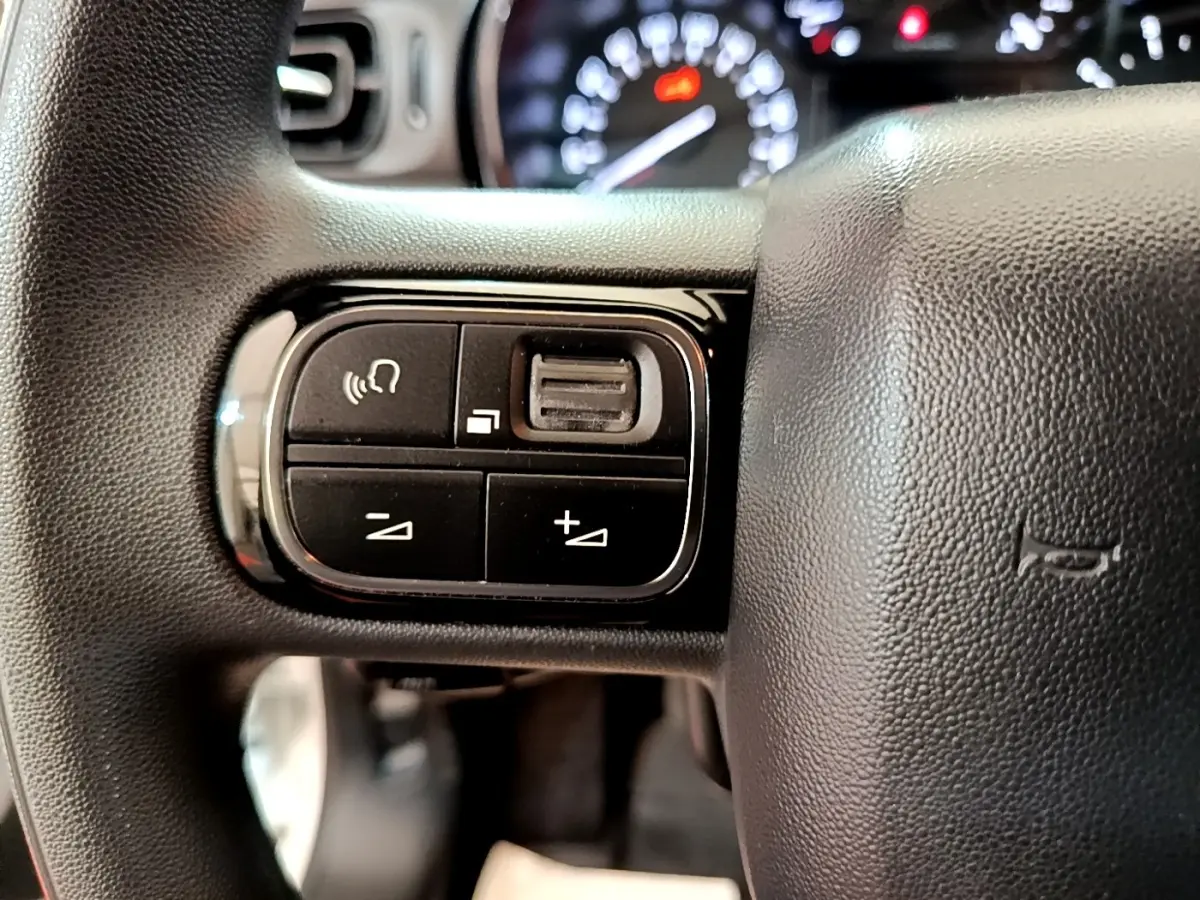 Gros plan sur les commandes de volume et téléphone au volant noir de la Citroën C3 blanche, tableau de bord flou en arrière-plan.