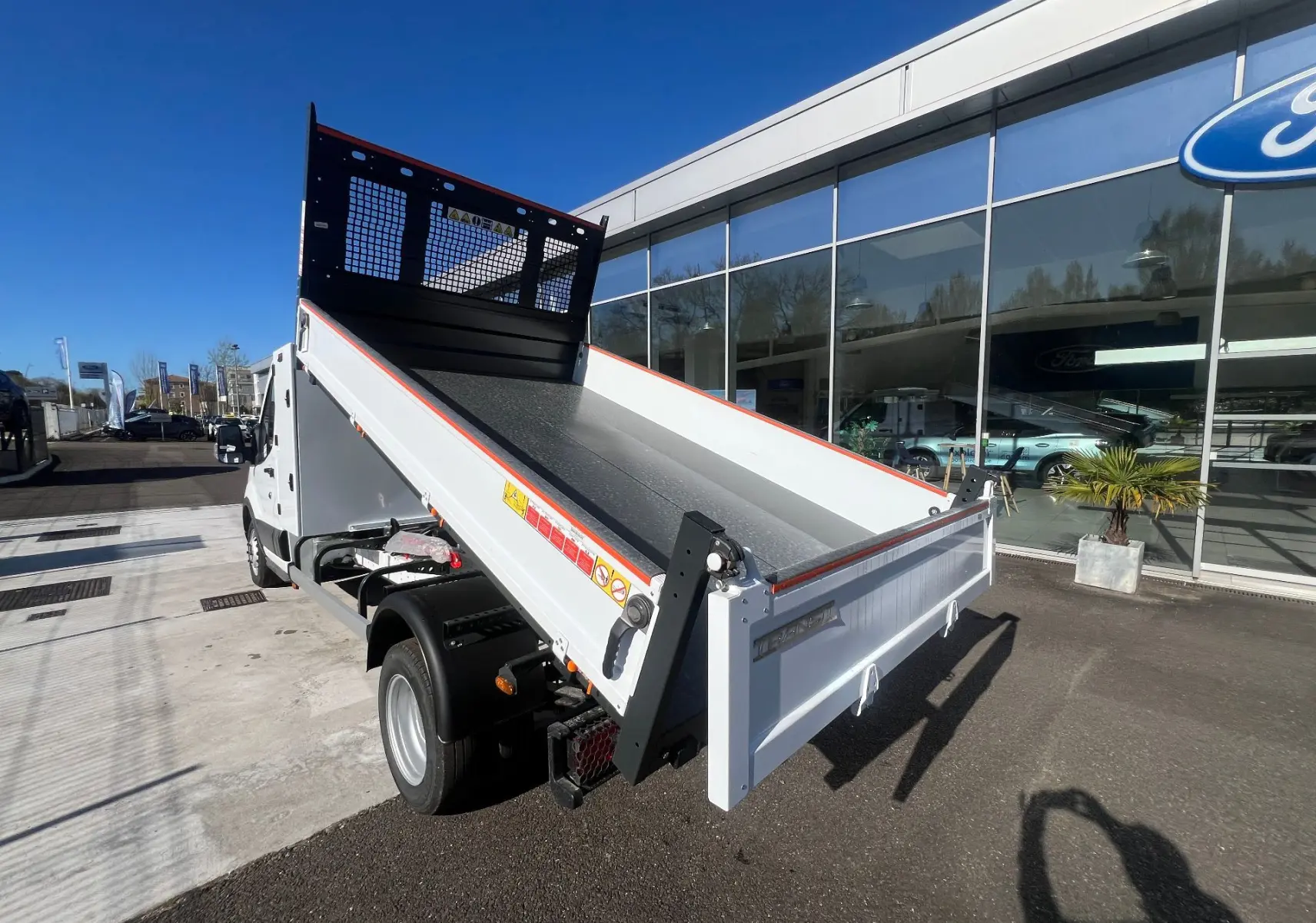 Vue 3/4 arrière droite du Ford Transit Chassis Cabine blanc avec benne relevée et coffre visible, stationné devant un bâtiment Ford.