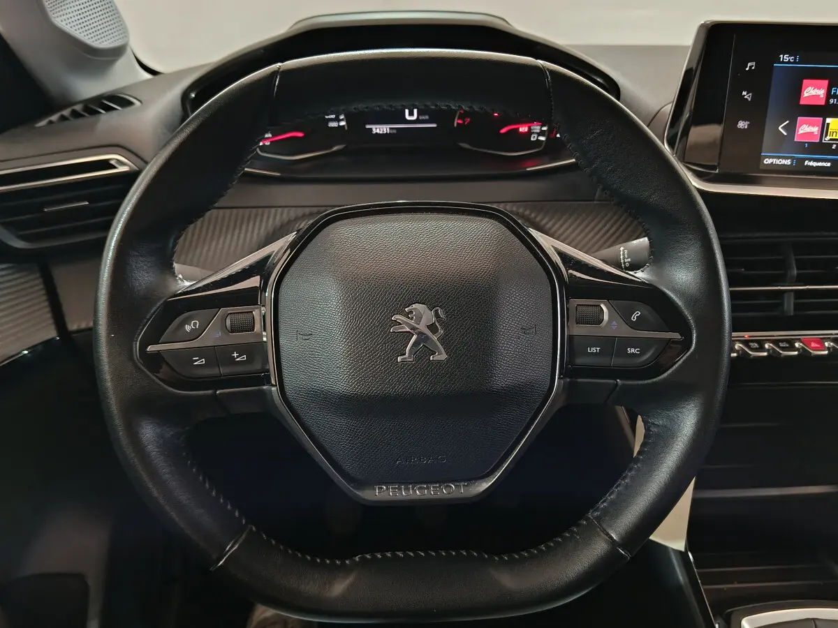 Vue rapprochée du volant cuir noir de la Peugeot 208 PureTech 100, avec tableau de bord digital et écran tactile visible.