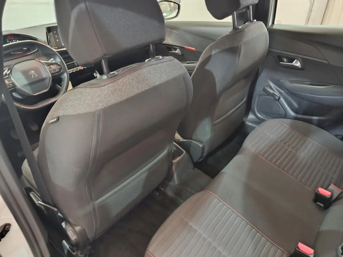Vue intérieure arrière de la Peugeot 208 blanche, montrant les sièges noirs avec surpiqûres rouges et le tableau de bord avant.