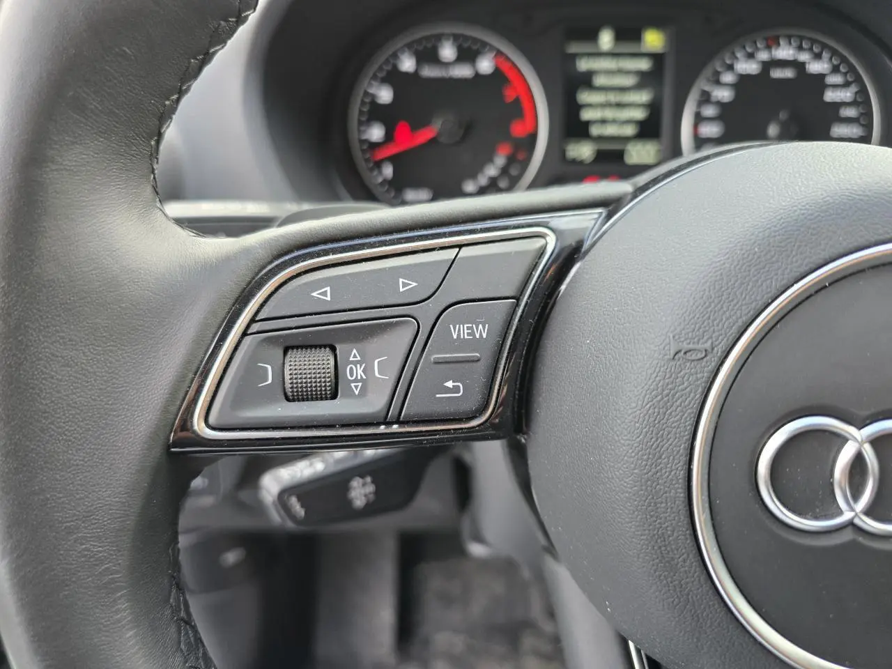 Gros plan sur les commandes à gauche du volant cuir d'un Audi Q2 noir mythic, avec compteurs en arrière-plan.