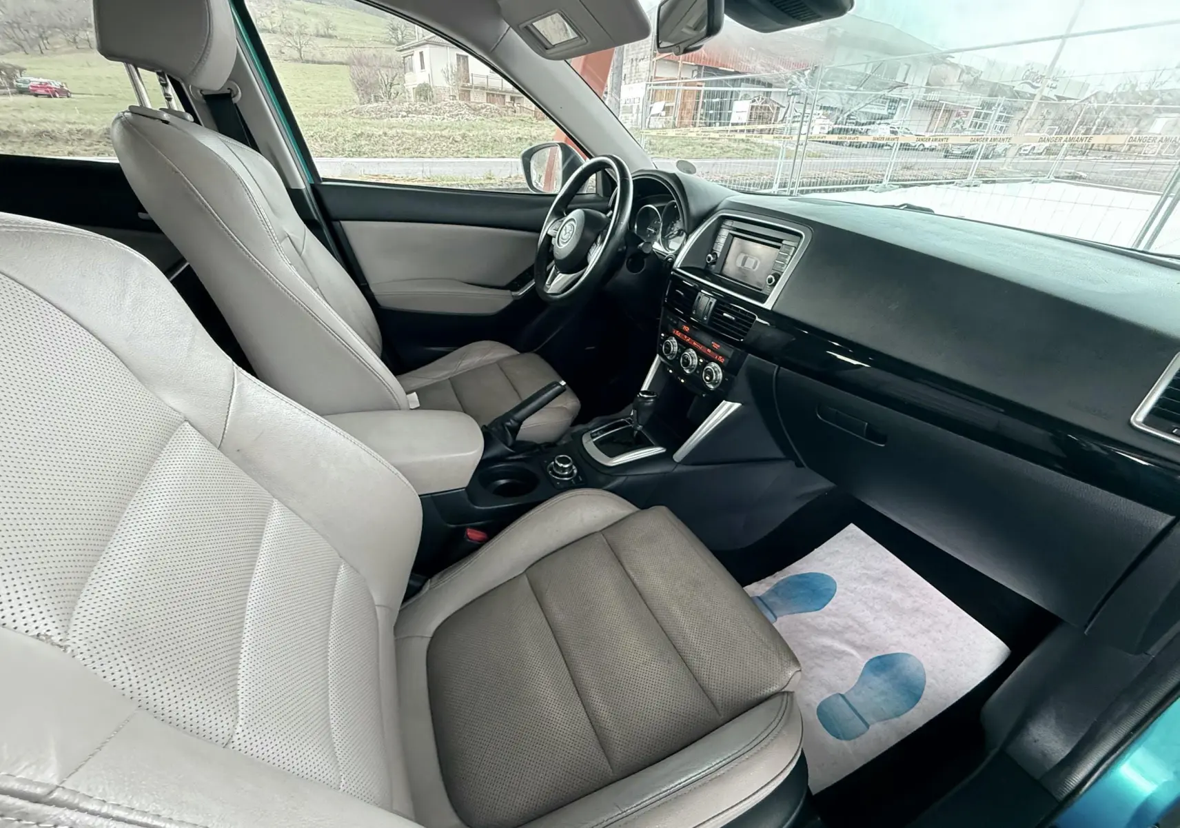 Vue intérieure côté conducteur du Mazda CX-5 bleu 2013, montrant sièges cuir beige et tableau de bord avec écran central.