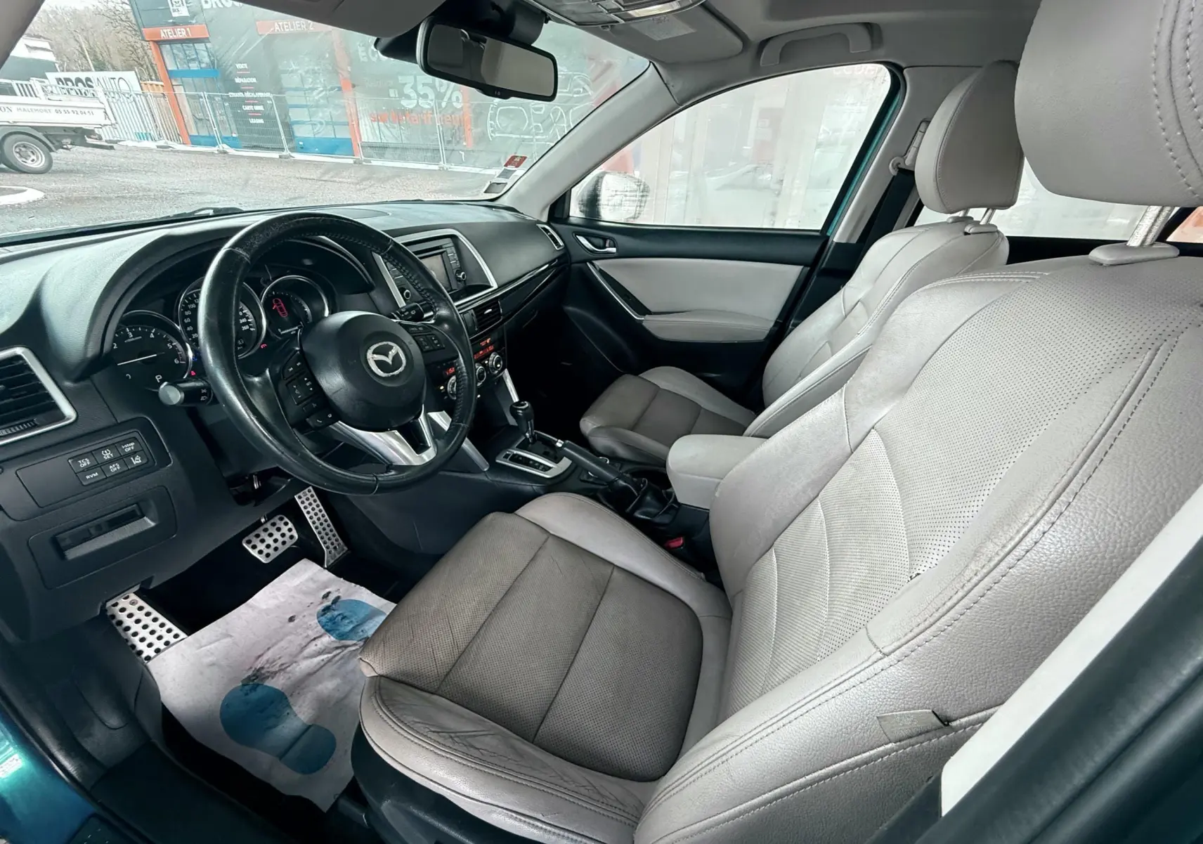 Vue intérieure côté conducteur du Mazda CX-5 bleu 2013, montrant les sièges en cuir clair et le tableau de bord avec volant multifonction.