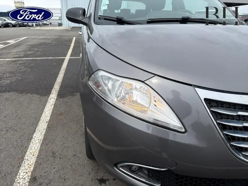Gros plan sur l'avant droit gris Argento de la Lancia Ypsilon 2014, mettant en valeur le phare et la calandre chromée.