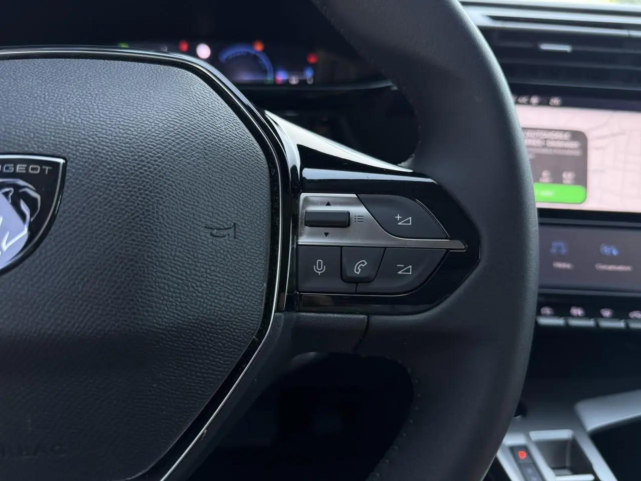 Gros plan sur le volant noir de la Peugeot 308 Hybrid 2025, avec commandes intégrées et écran tactile de navigation en arrière-plan.