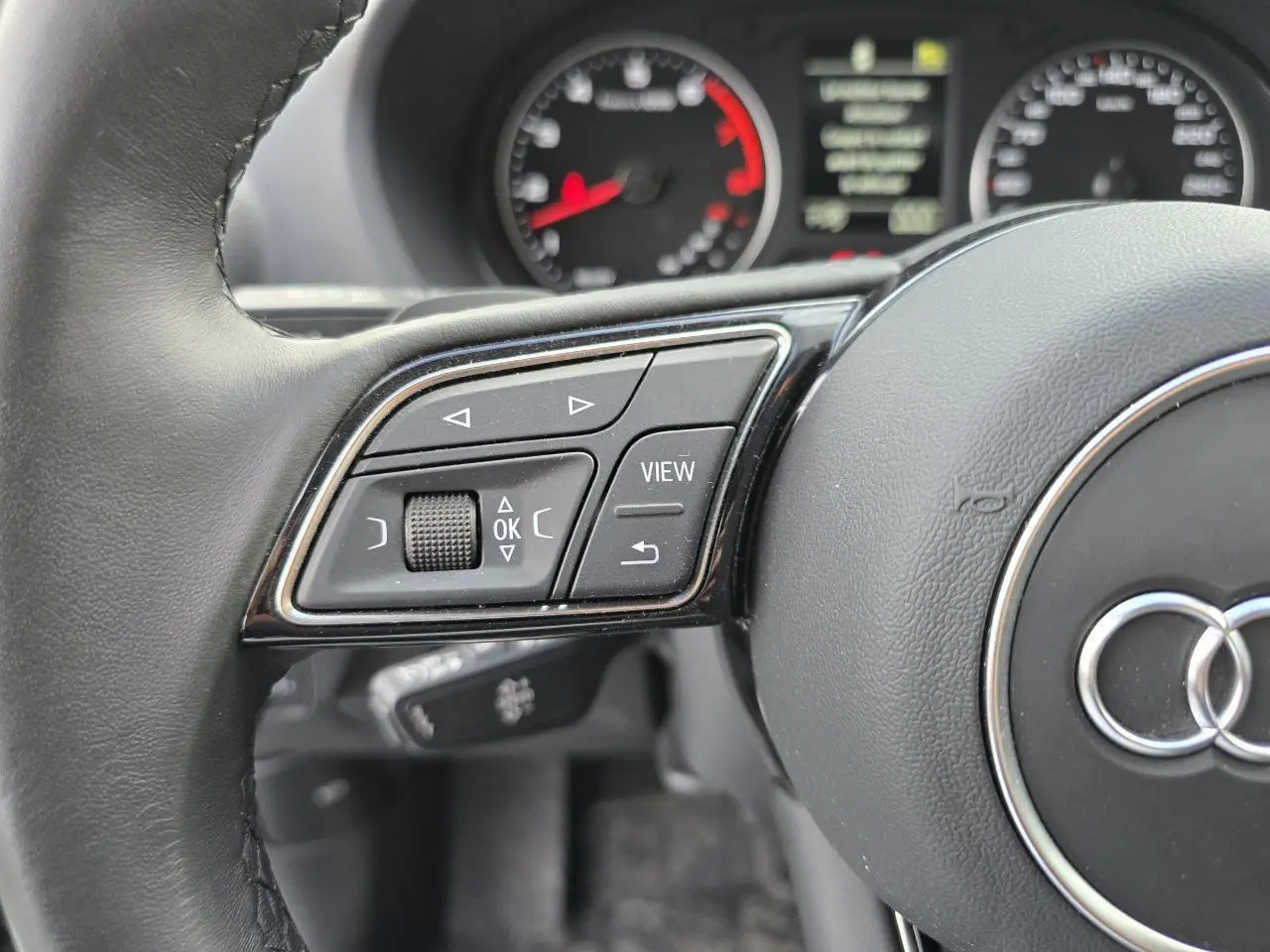 Gros plan sur la partie gauche du volant cuir noir de l'Audi Q2 2023 avec commandes multifonctions et tableau de bord flou.