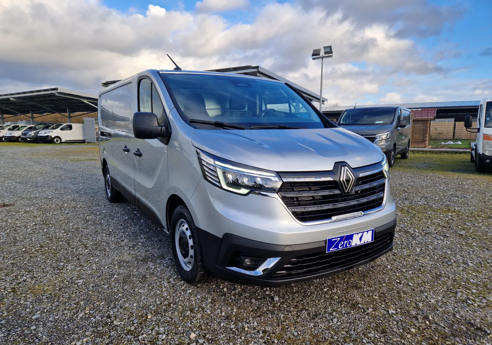Vue 3/4 avant droit d'un Renault Trafic Fourgon gris Highland avec calandre noire et feux LED allumés sur un parking.